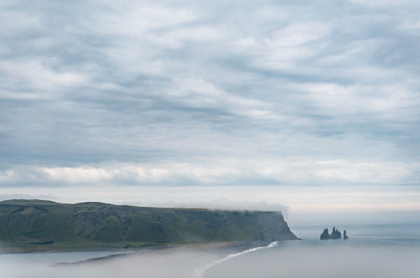 Coastal landscape with dramatic cliffs under a cloudy sky, partially veiled in mist. The scene conveys a serene and mysterious atmosphere.
