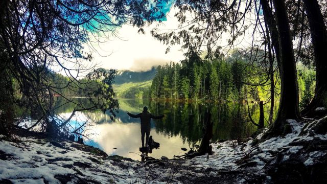 Man stands with open arms on a snowy lake shore, surrounded by tall trees. Reflective water mirrors a lush forest and cloudy sky, evoking tranquility.
