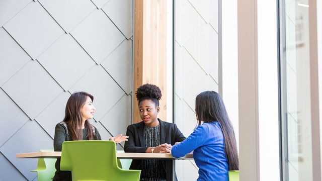 A culturally diverse group of 3 female Royal Roads University students sit together in green chairs around a table having a conversation in one of the schools modern campus buildings.