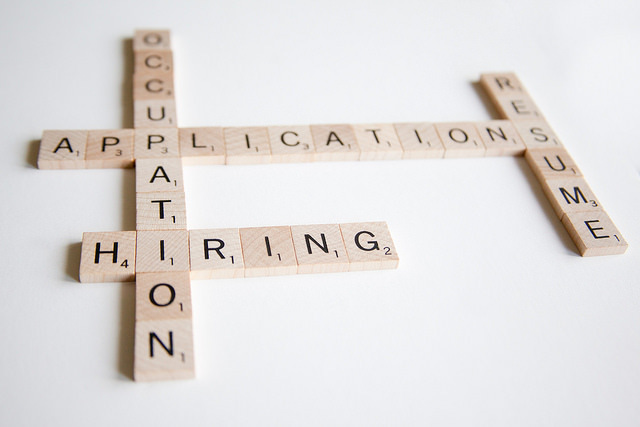 Scrabble tiles spell "Applications," "Resume," "Hiring," and "Occupation" in a crossword layout on a white surface, conveying a job-related theme.