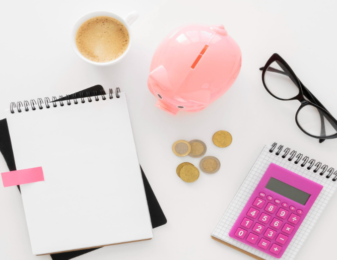 A flat lay of a pink piggy bank, coffee cup, notebooks, coins, a pink calculator, and black glasses on a white surface.