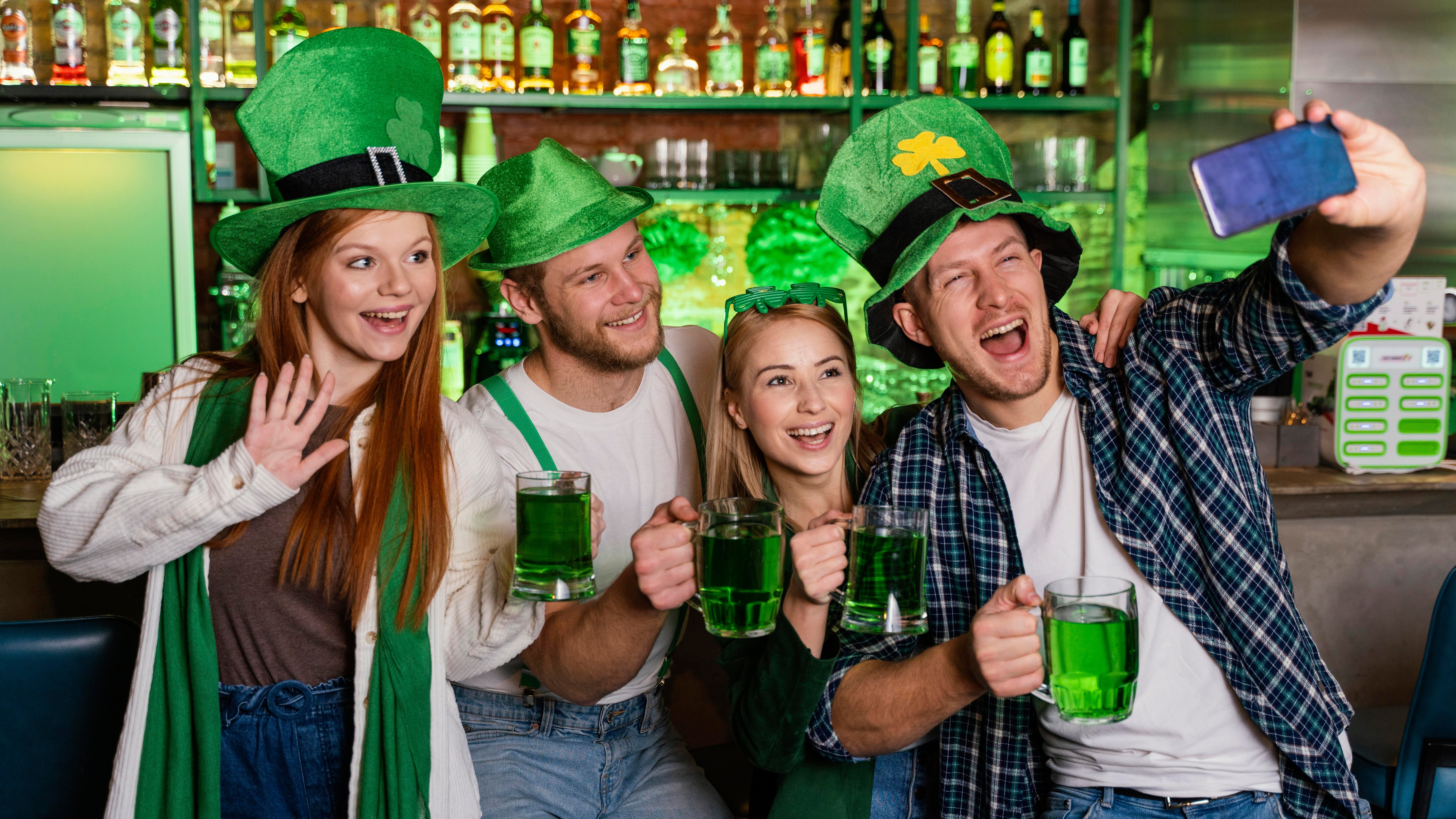 Four people celebrate St. Patrick's Day in a bar, wearing green hats and holding mugs of green beer, smiling and taking a selfie. Festive and joyful mood.