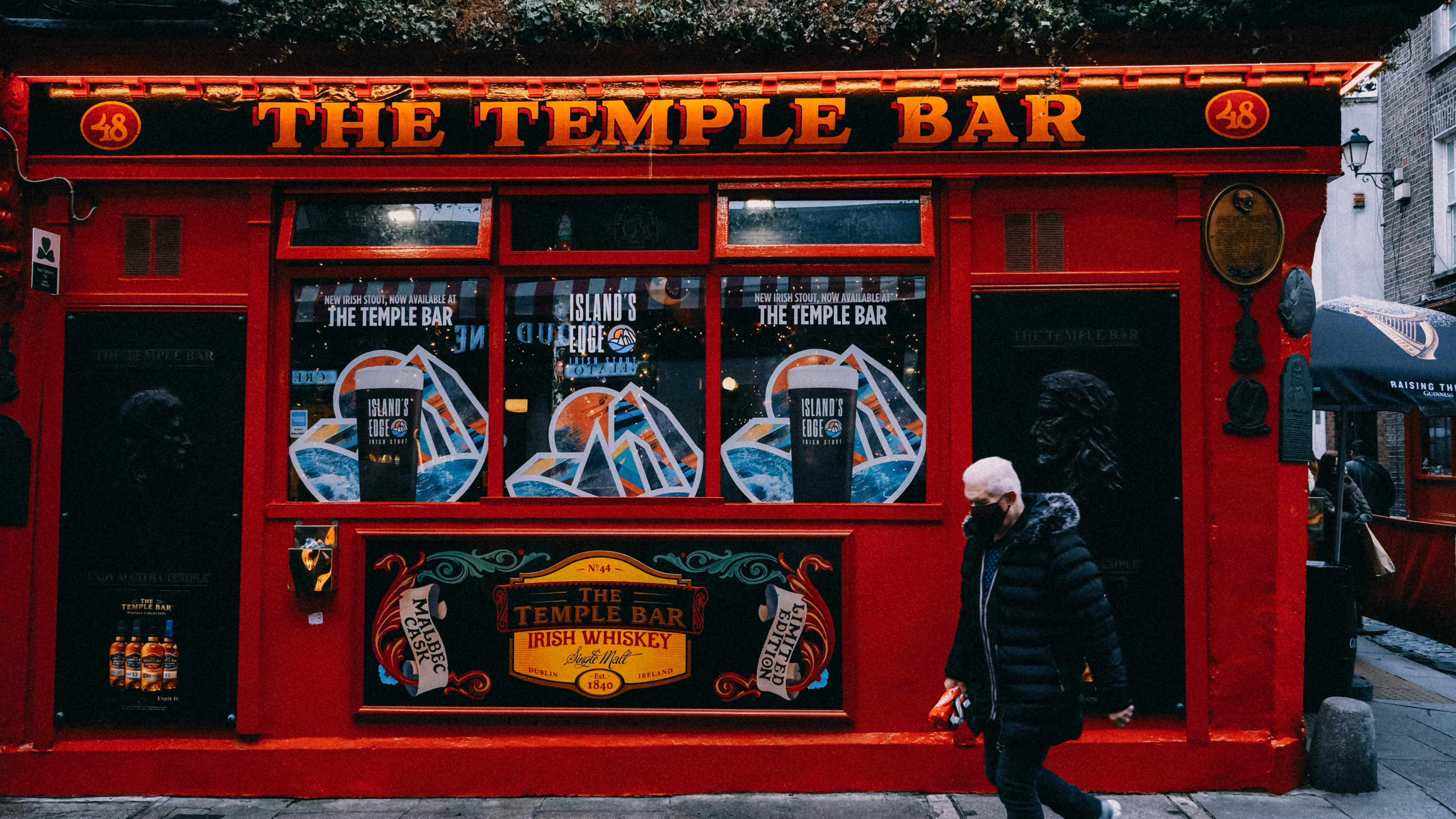 A person walks past the iconic red façade of The Temple Bar pub adorned with neon signs and posters. The scene conveys a lively, inviting atmosphere.