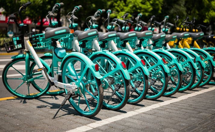 : A line of turquoise rental bicycles is neatly parked on a sunny street, with trees in the background. The mood is bright and orderly.