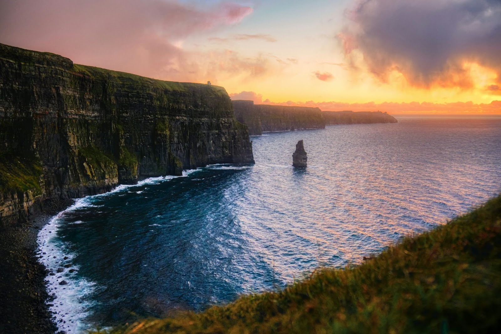 Sunset over the Cliffs of Moher, with dramatic cliffs meeting the ocean. A lone sea stack rises from the water. Sky glows with warm hues.
