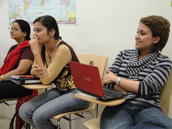 Three women sit attentively in a classroom with notebooks and a laptop. A world map is on the wall, suggesting a learning environment.