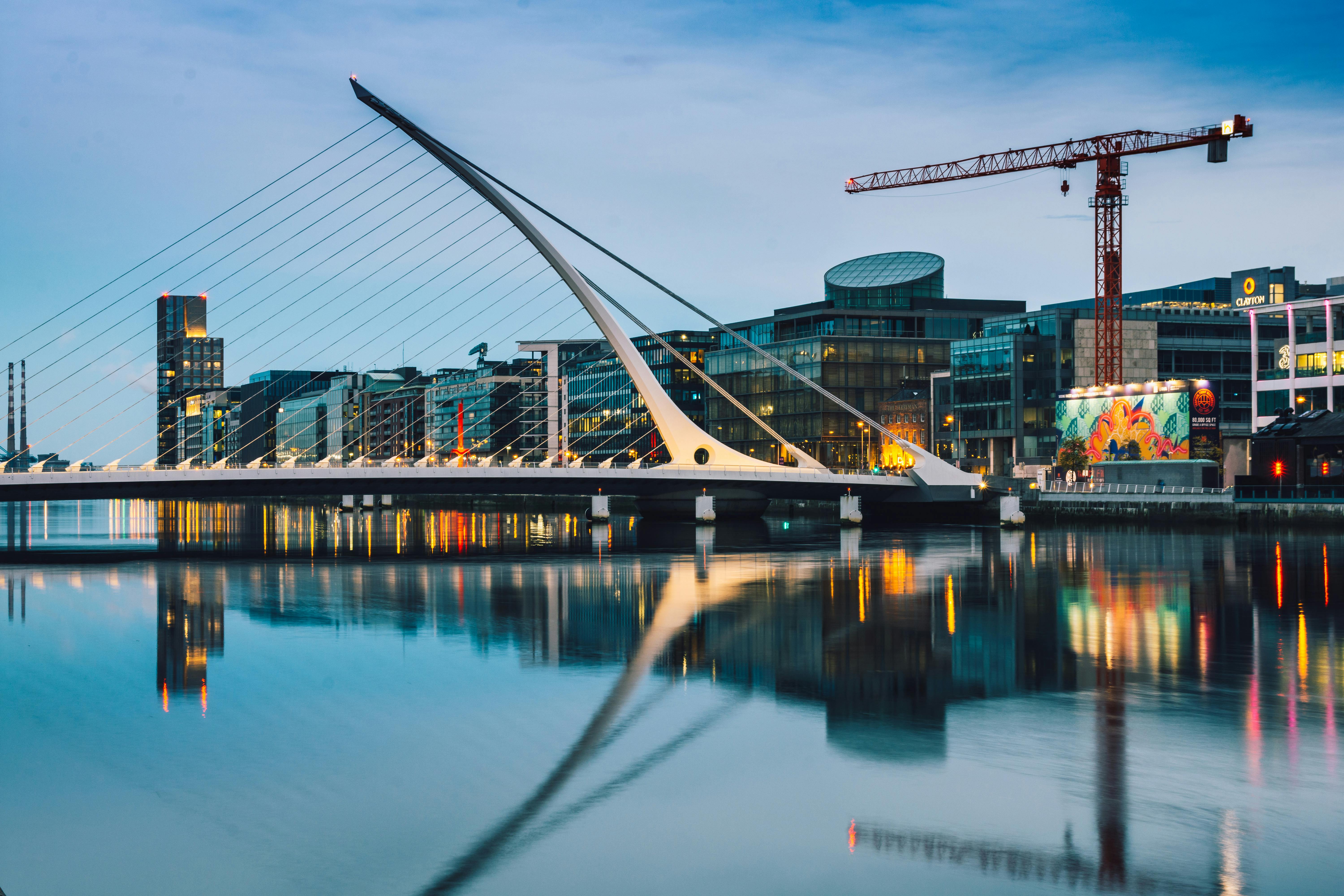 A sleek, modern bridge with harp-like cables spans a calm river, reflecting city lights. The backdrop features contemporary buildings and a crane, evoking urban development.