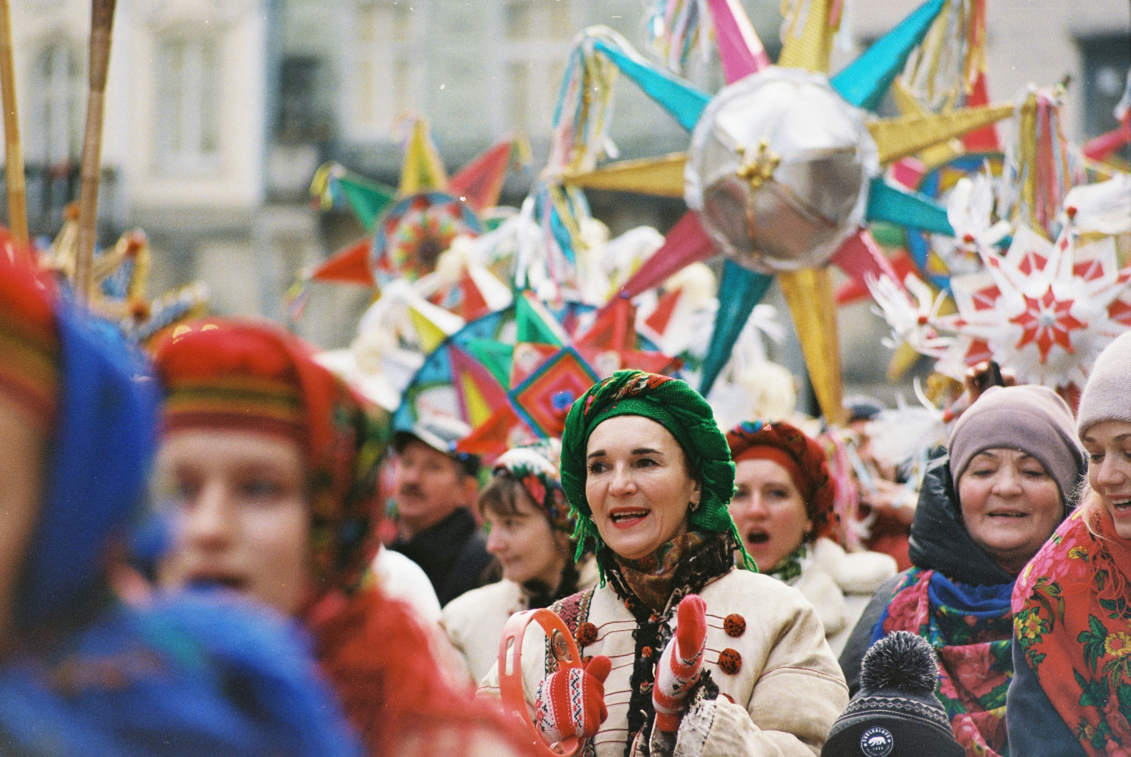 A festive crowd in colorful traditional clothing celebrates with large, vibrant star decorations. The atmosphere is joyous, lively, and communal.