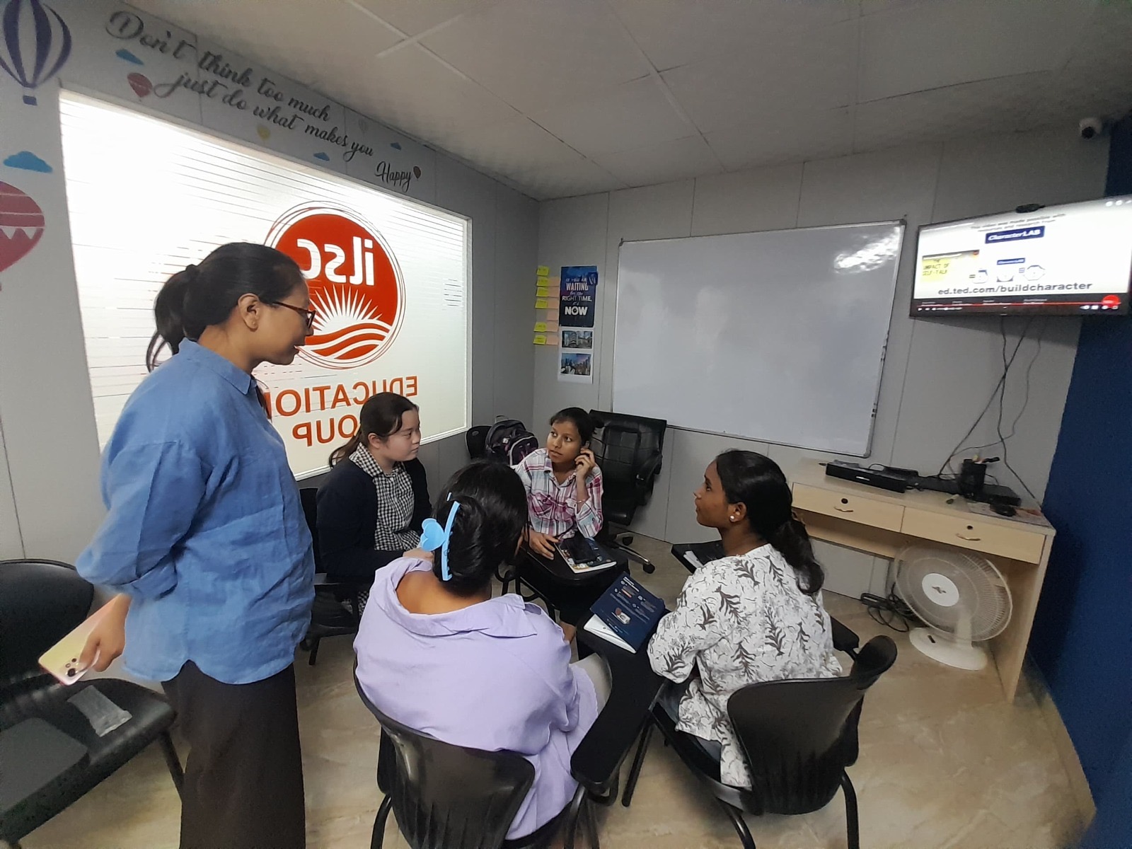 A group of five people in a classroom engage in discussion. They sit on chairs around a laptop. A whiteboard and wall-mounted screen are visible.