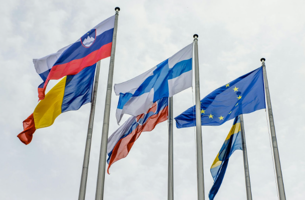 Several flags flutter against a cloudy sky, including those of Slovenia, Finland, Sweden, Russia, Romania, and the European Union.