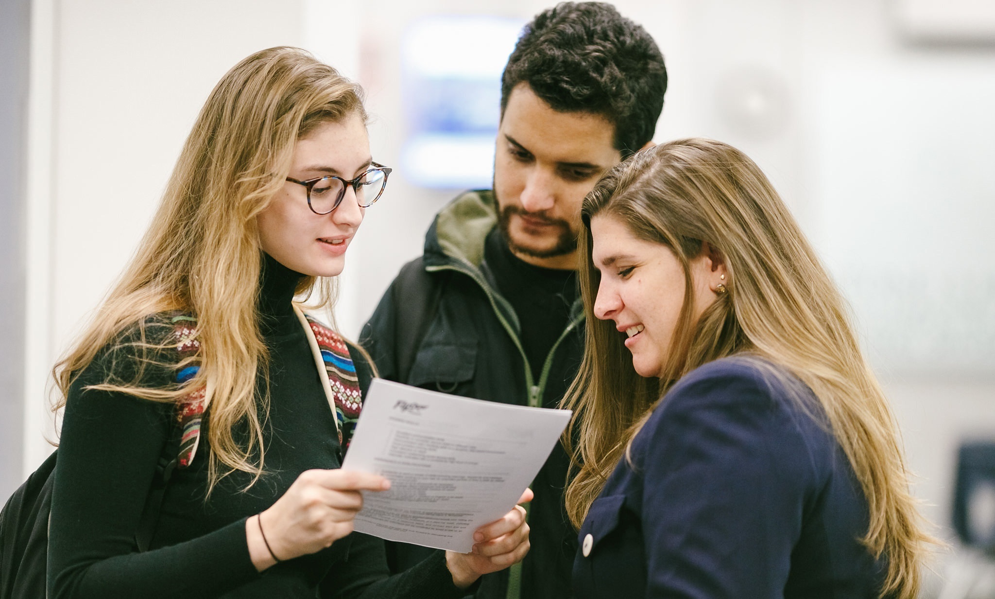Three people stand closely together, focused on a document one of them holds. They appear engaged and collaborative, in a casual setting.