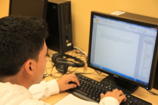 A person in a white shirt is working on a desktop computer, focused on typing a document. The setting appears to be a computer lab with a calm atmosphere.