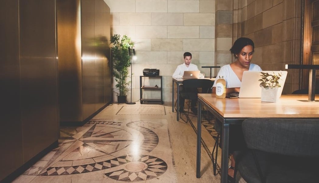 Two people work quietly on laptops in a stylish, modern office with patterned flooring and soft lighting. A small potted plant and a drink are on the table.