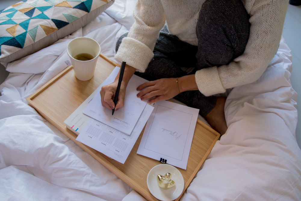 Person in cozy sweater writing on papers on a wooden tray with coffee, seated on a bed. Atmosphere is calm and focused, suggesting planning or journaling.