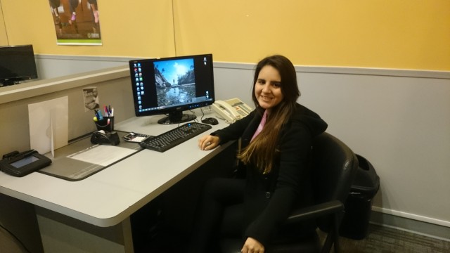 A smiling woman sits at an office desk with a computer, phone, and stationery. She appears relaxed in a brightly lit, yellow-walled room.