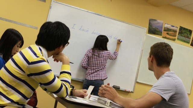 A classroom setting with a woman in a plaid shirt writing on a whiteboard. Three students sit at desks, attentively focused on the lesson. The atmosphere is studious.