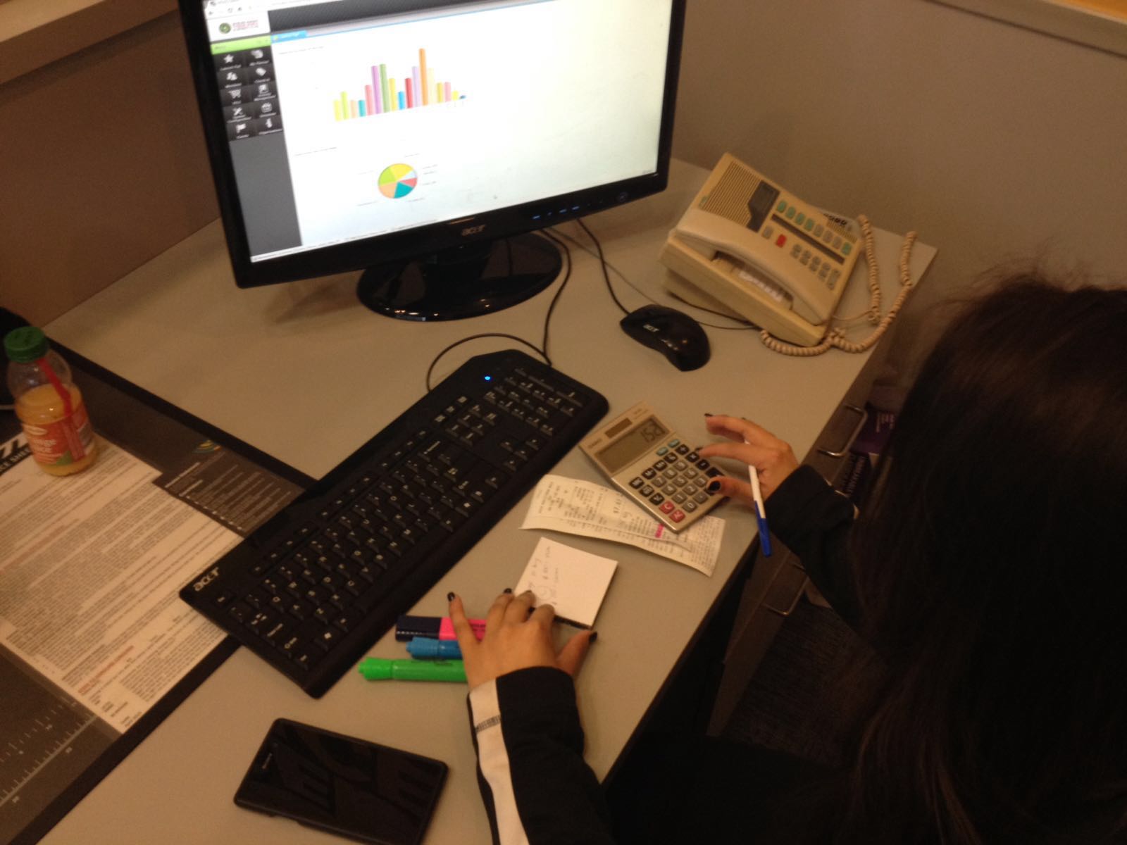 A person works at a cluttered desk with a computer displaying charts, using a calculator and taking notes. The scene conveys focus and diligence.