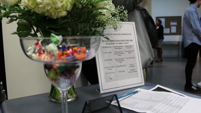 A career fair table with a glass bowl of colorful candy, a floral arrangement, and papers. A sign lists employer partners. The setting is busy and professional.