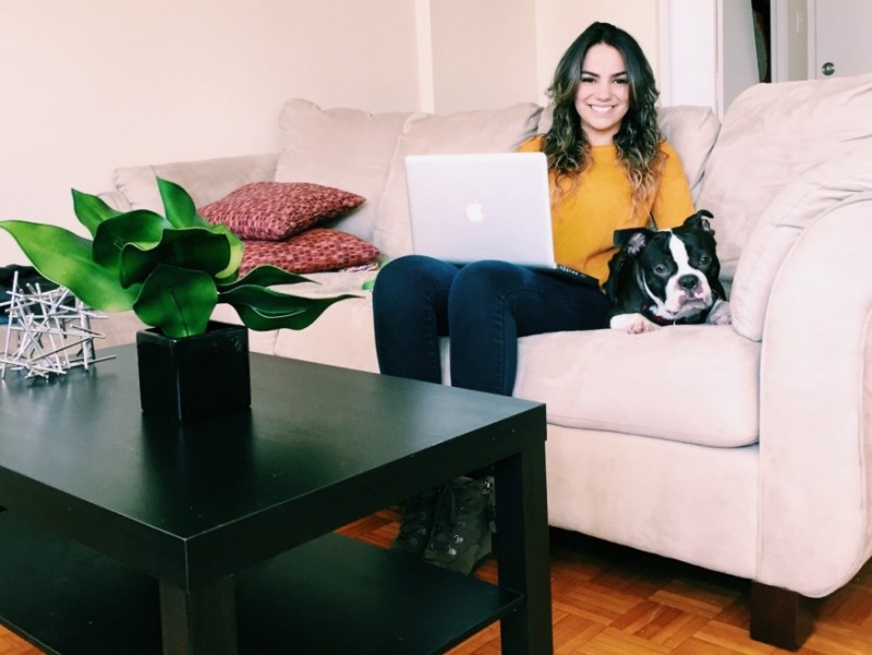 A woman in a yellow sweater sits on a beige sofa with a laptop, a black and white dog beside her. A coffee table with a green plant is in the foreground.