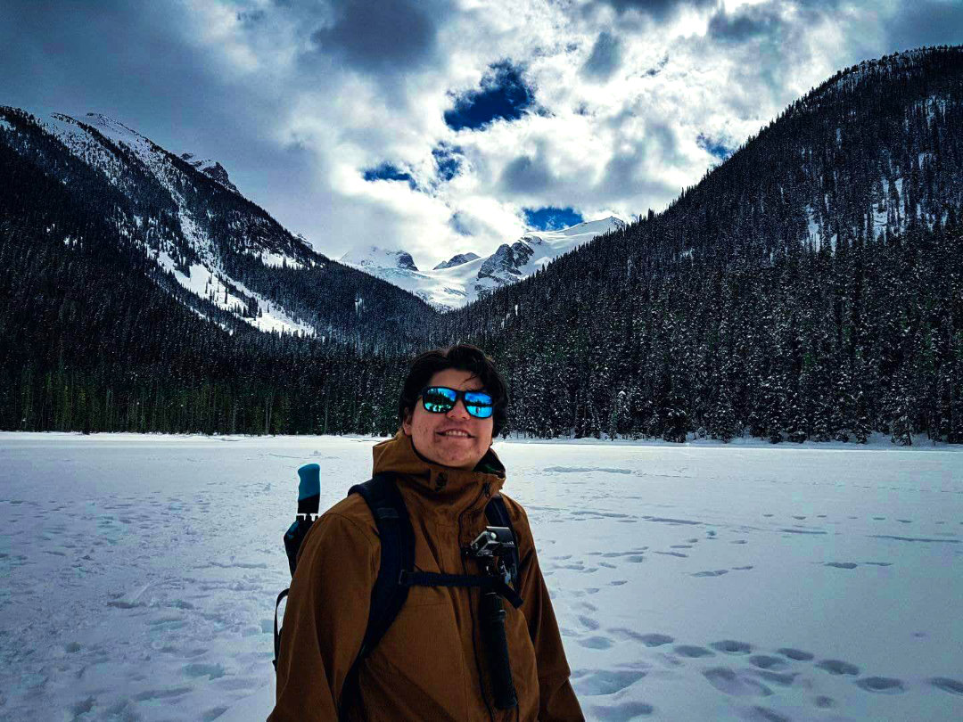A person in sunglasses smiles, standing on a snow-covered landscape with snow-draped mountains and a cloudy blue sky in the background, exuding a sense of adventure.