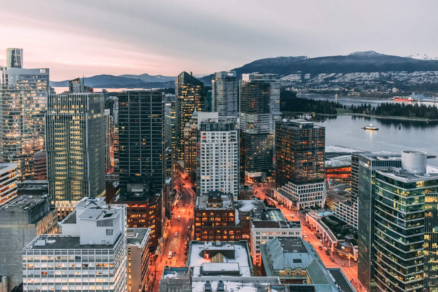 Aerial view of a bustling cityscape at dusk, featuring illuminated skyscrapers and streets. In the background, calm waters and mountains are visible under a soft sky.