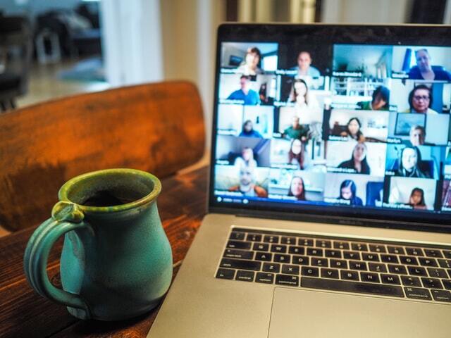 A laptop displaying a Zoom meeting with many participants, beside a green ceramic mug on a wooden table, conveying a cozy remote work setting.