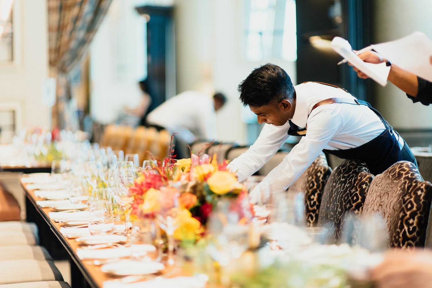 A waiter in a white shirt and black apron sets a table in a formal dining room. Vibrant flowers and neatly arranged glassware create an elegant atmosphere.