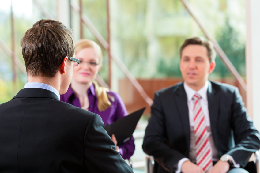 Three people in a bright office setting, engaged in a business meeting. Two face the camera with attentive expressions, while one has their back turned.