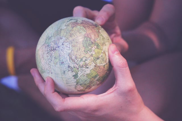 Two hands gently cradle a small globe, depicting continents and countries. The image conveys care and curiosity, with a softly blurred background.