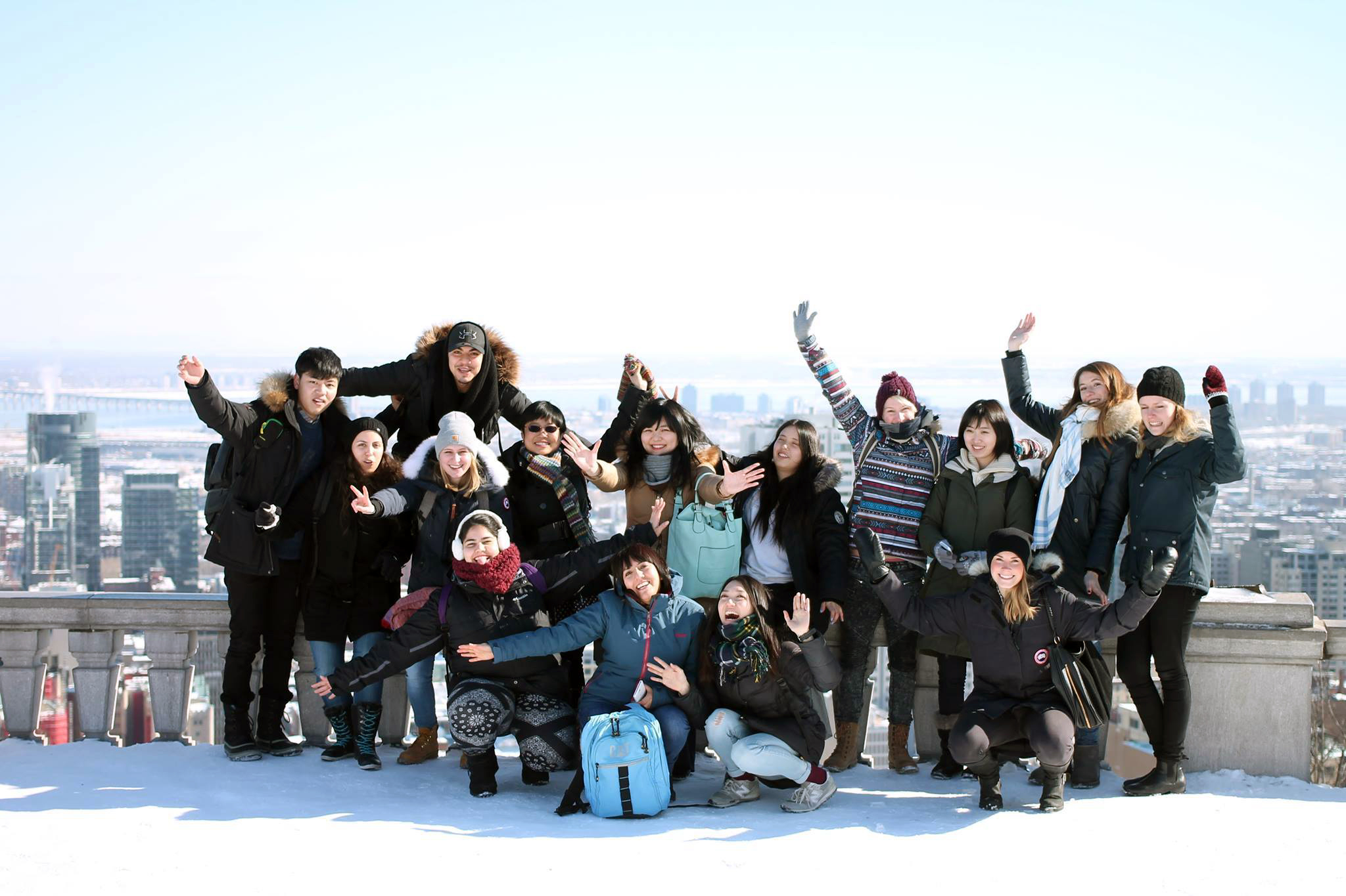 A joyful group of people pose on a snowy city rooftop, arms raised in celebration. The bright sky and distant skyline create a lively, energetic mood.