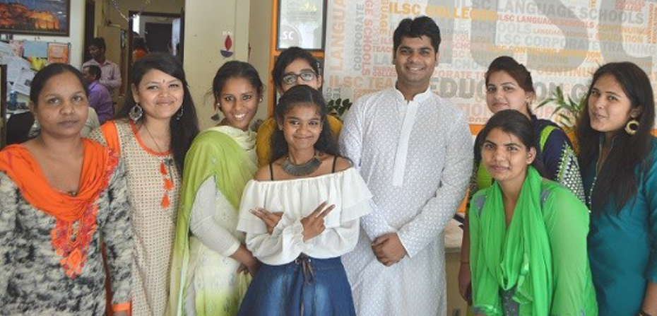 A group of nine people smiling and standing together, wearing colorful traditional and casual attire in a cheerful, friendly setting.