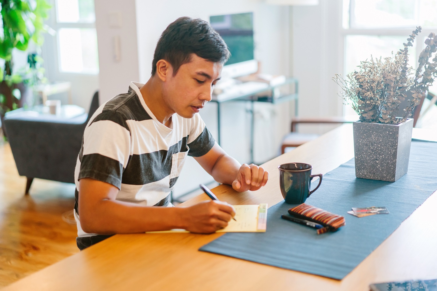 A young man in a striped shirt writes on a notepad at a wooden table. A coffee mug, pens, and a potted plant are nearby, creating a focused, calm atmosphere.