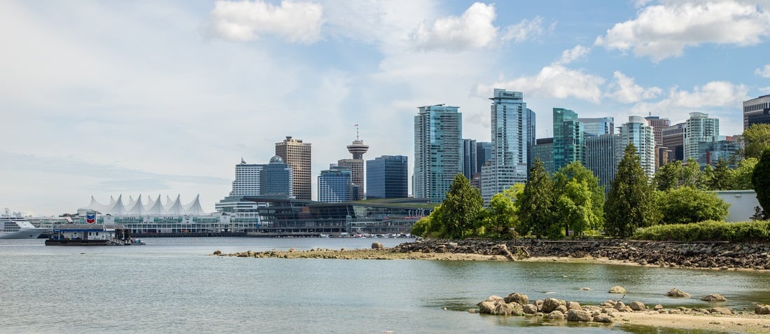 Skyline of a city with modern skyscrapers and a distinctive sail-like building by the waterfront. Calm waters and a rocky shore in the foreground.