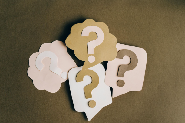 Four paper cut-outs with question marks on speech and thought bubbles in pink, beige, and white, arranged on a brown background, convey curiosity.