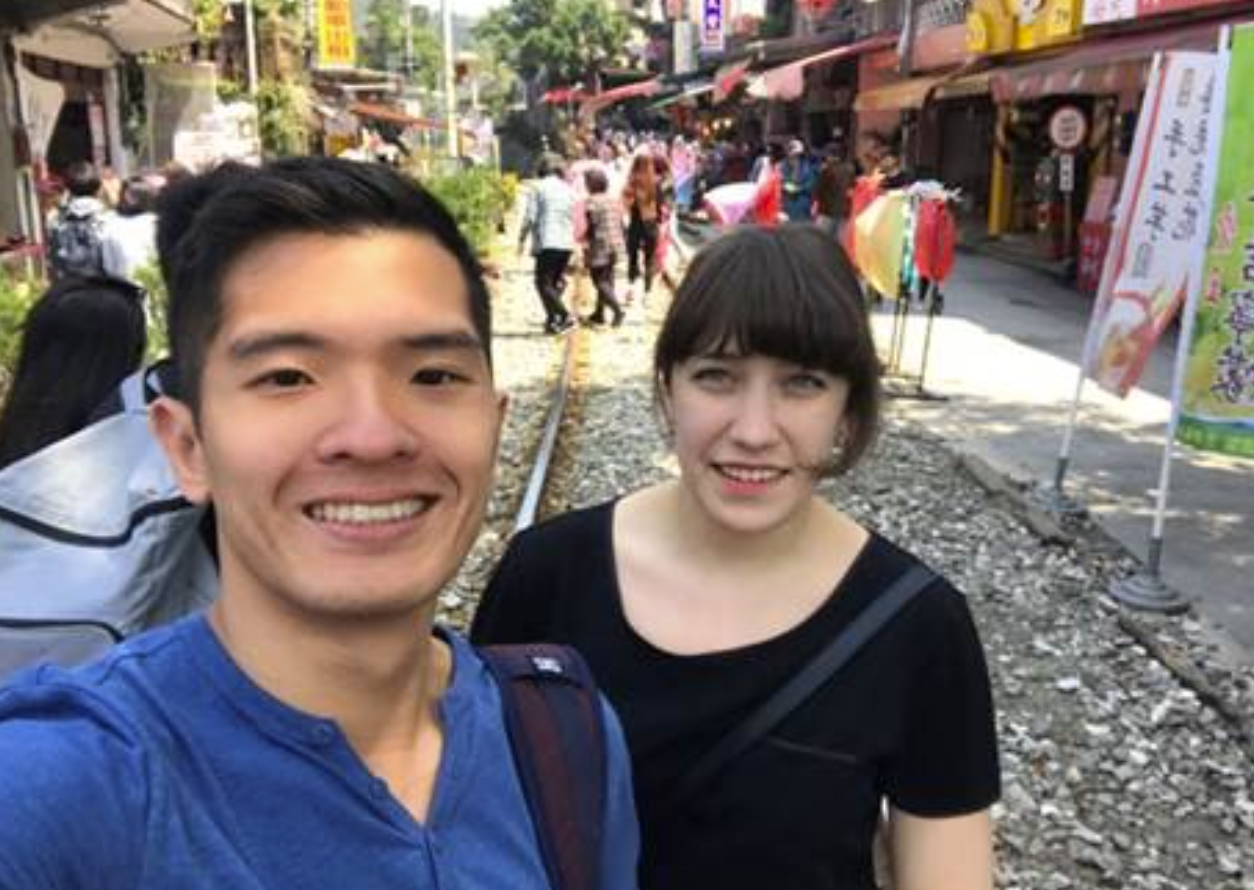 A smiling man and woman take a selfie on a cobblestone path with train tracks. The background features bustling shops, colorful signs, and people.