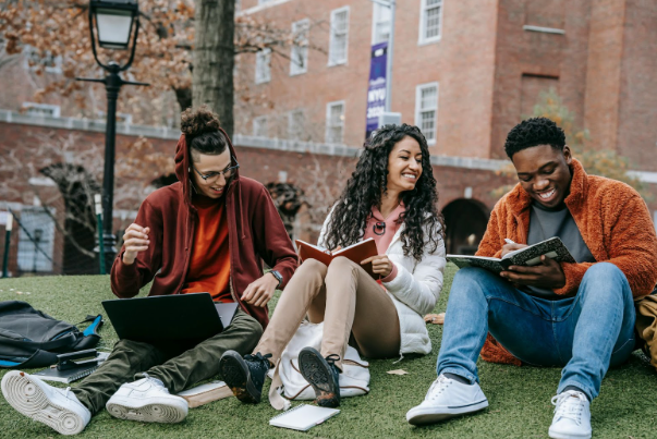 Three friends sit on grass, laughing and studying outside a brick building. One has a laptop, another a notebook, and all appear engaged and happy.
