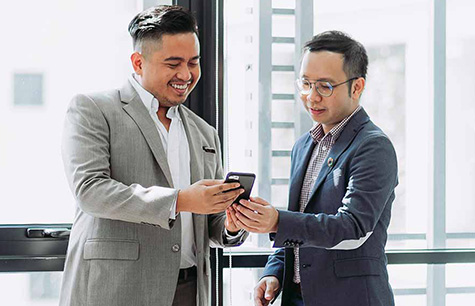 Two male colleagues in an office discussing something on a cell phone.  Take a practicum program at Greystone College Montreal to get unpaid work experience