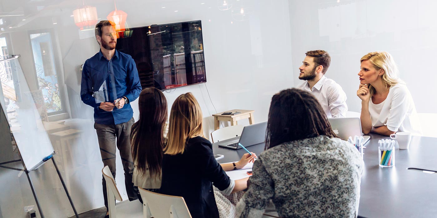 Office setting, male giving presentation to 4 women colleagues, and 1 male colleague.  Study business at Greystone College career college Canada in Montreal, Toronto or Vancouver.