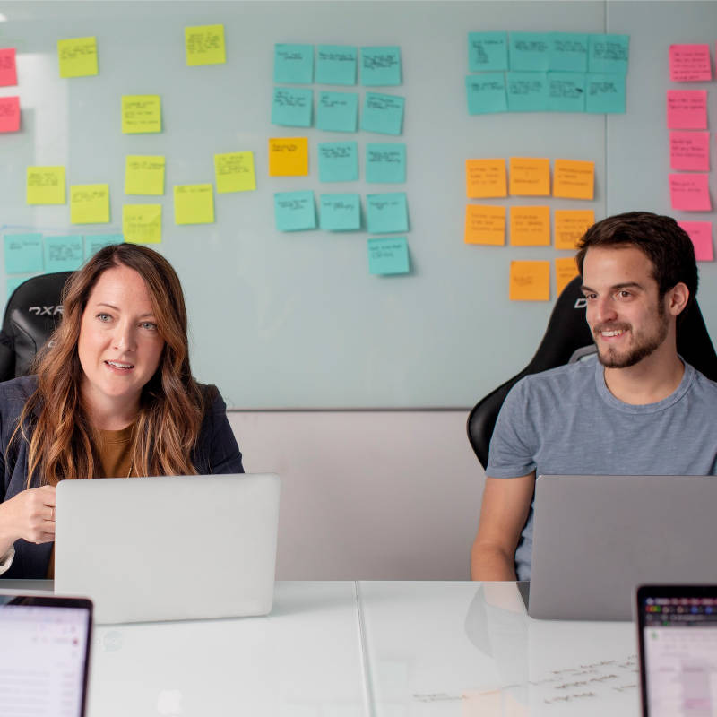 two colleagues one female one male sit in front of laptops in a meeting with coloured sticky notes on a white board behind them - study leadership and innovation in Australia