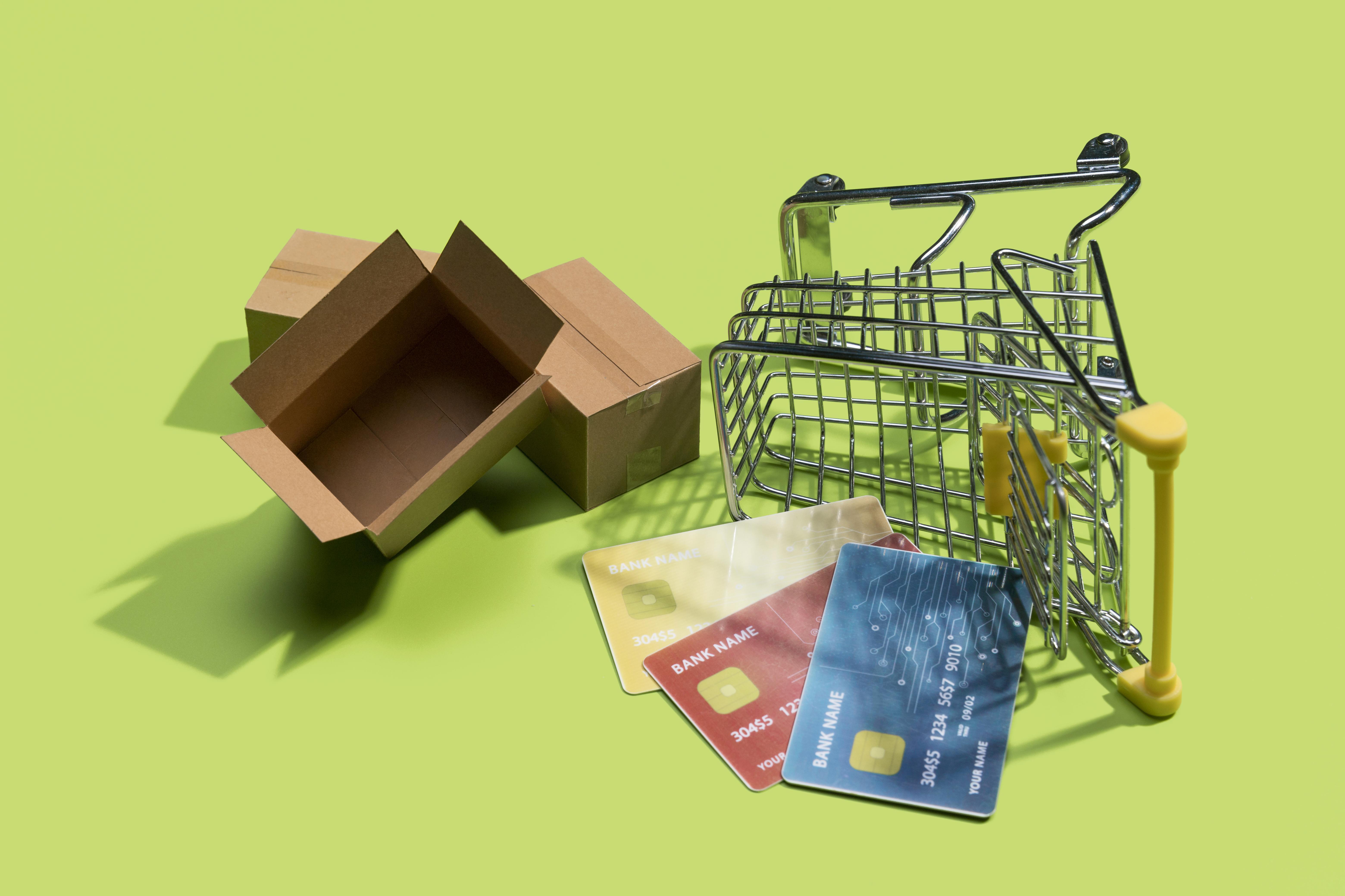 A small shopping cart tipped over next to an open cardboard box and three credit cards on a bright green background, suggesting online shopping.